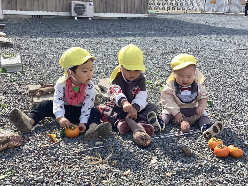 いもほりで収穫したさつまいもと、地域の方からいただいた柿を囲んで座っている3人の園児。