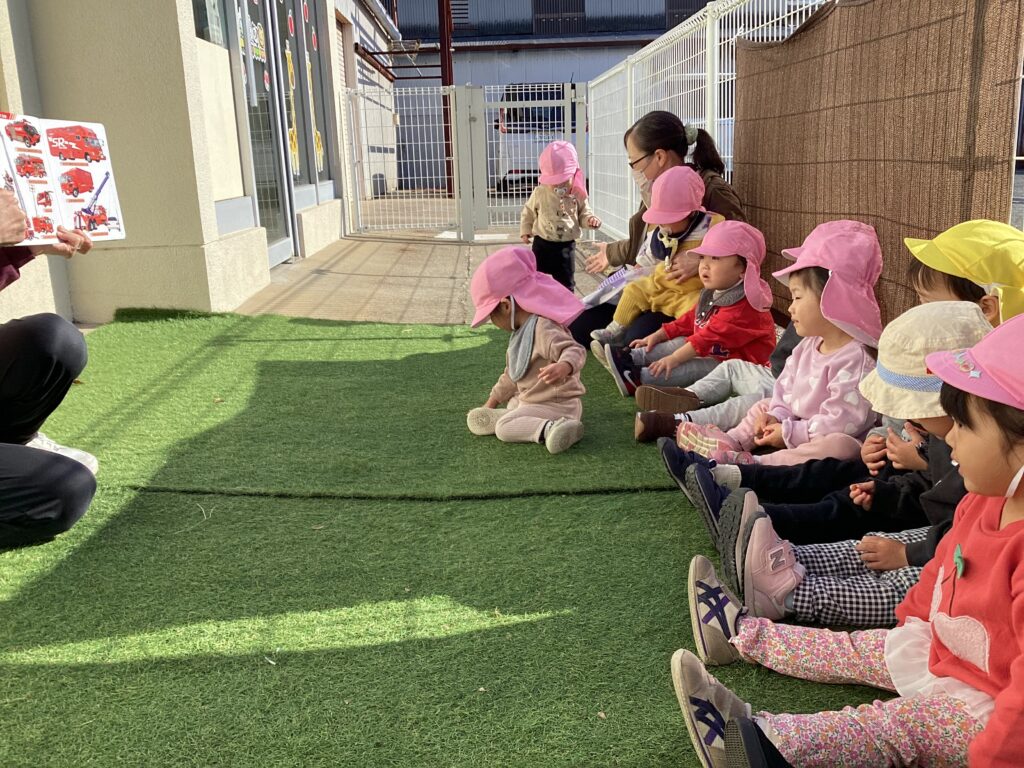 保育士が消防車の絵本を見せ、水消火訓練の前に説明を聞くタイムカプセル保育園の園児たち。