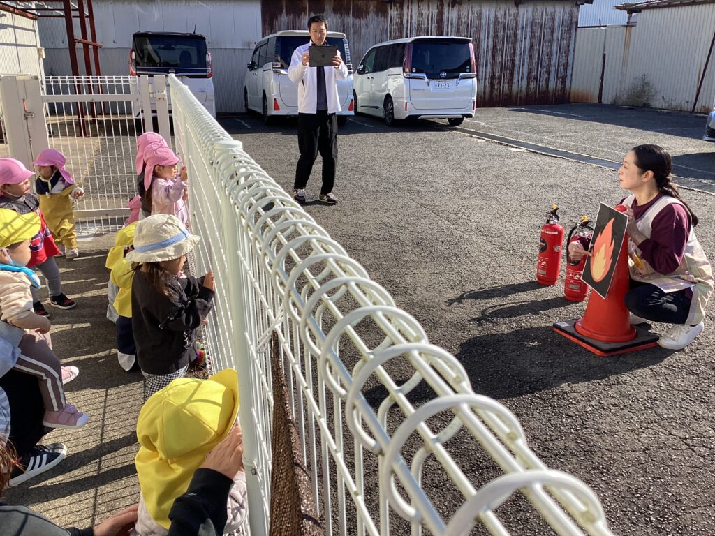 水消火訓練で、保育士が消火器と炎のパネルを使って説明する様子をフェンス越しに見学するタイムカプセル保育園の子どもたち。