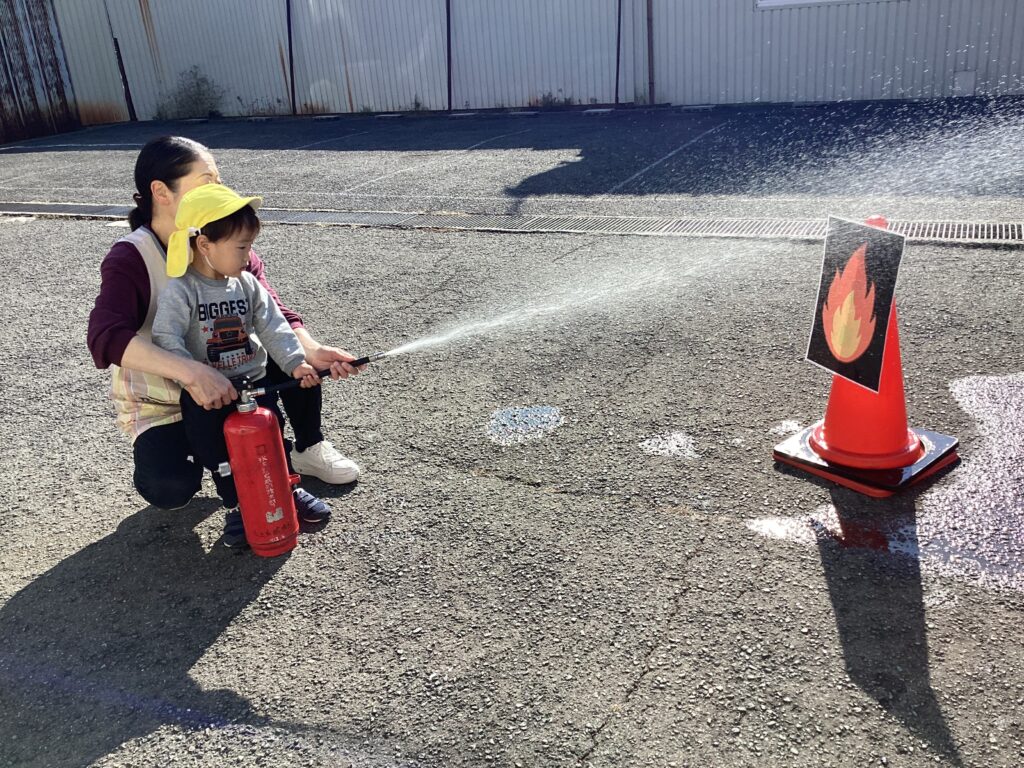 タイムカプセル保育園の避難・消火訓練。先生に補助されながら消火器を操作し、安全への意識を学ぶ園児。