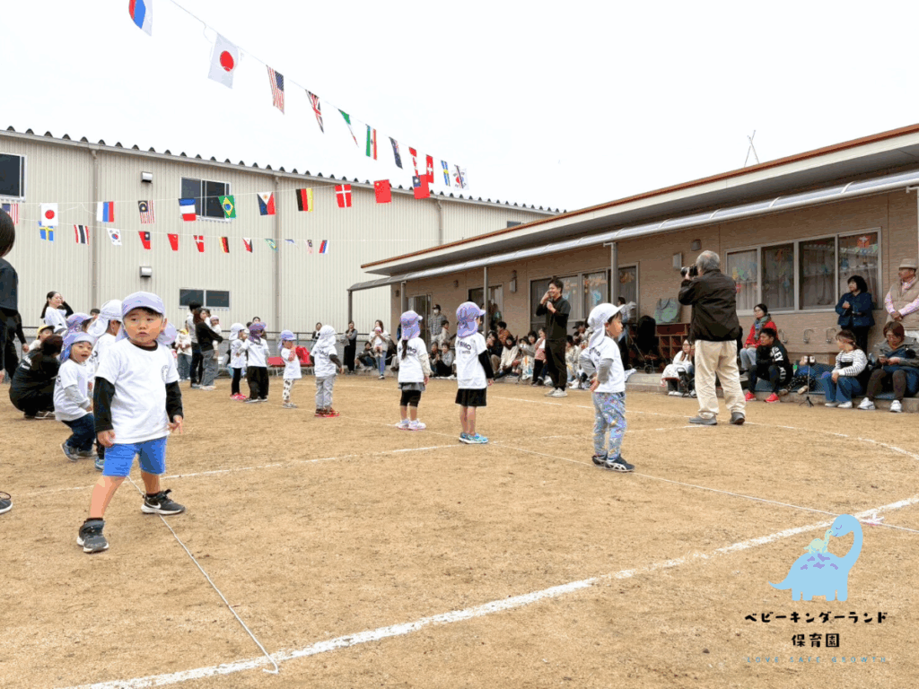 タイムカプセル保育園の運動会のグラウンドにて、白いTシャツと薄い紫の帽子をかぶった園児たちが、建物の前で整列している。