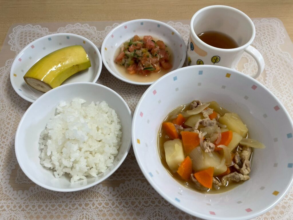 保育園の献立例。メインは具材が大きくカットされた肉じゃが、白いご飯、デザートには皮をむいたバナナ、そしてトマトとキュウリの和え物のような副菜と飲み物（お茶）。風邪予防にもなる栄養満点で彩りの良い給食の様子が伝わる。