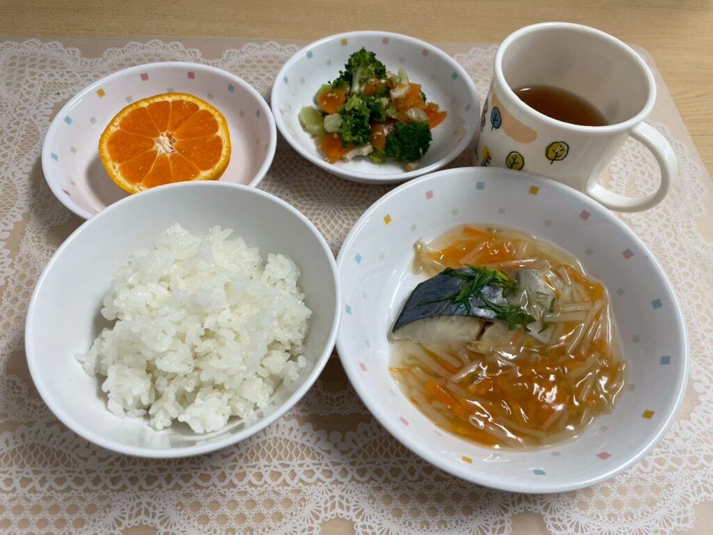 保育園の給食の献立例。メインは、切り身のサワラに、もやしや人参がたっぷり入った餡がかかった「さわらのもやしあんかけ」。ご飯、ブロッコリーなどが入った副菜、カットされたみかん、飲み物が添えられた、彩り豊かで栄養バランスの取れた和食の献立。