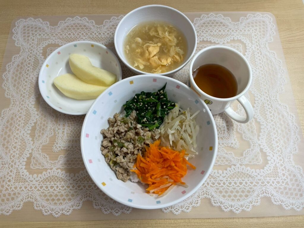 保育園の給食の献立例。ご飯の上に、そぼろ肉、人参の細切り、茹でたもやしやほうれん草などが彩りよく乗った「三色どんぶり」（またはビビンバ風どんぶり）。卵スープと、皮をむいたカットりんご、お茶が添えられた、栄養バランスの良い献立。