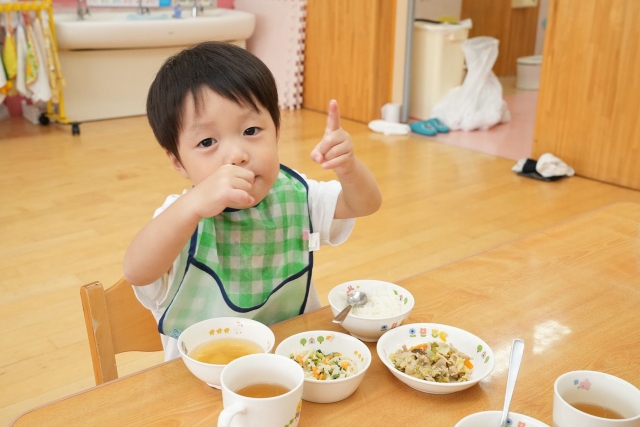保育園の給食の時間。チェック柄のエプロンをつけた幼い男の子が、小さなテーブルと椅子に座り、ご飯や汁物など複数の小皿が並んだ給食を食べている。