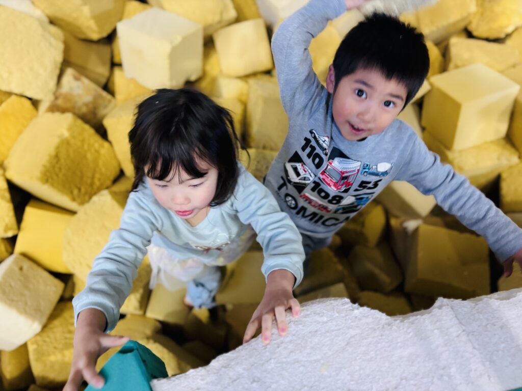 兵庫県丹波市のディナモ体操クラブ体育館にて、スポンジプールの縁を一生懸命に登るタイムカプセル保育園の子どもたちのクローズアップ写真。黄色いスポンジの中から顔を出し、真剣な表情や好奇心いっぱいの瞳でお掃除や遊びを楽しんでいる様子。