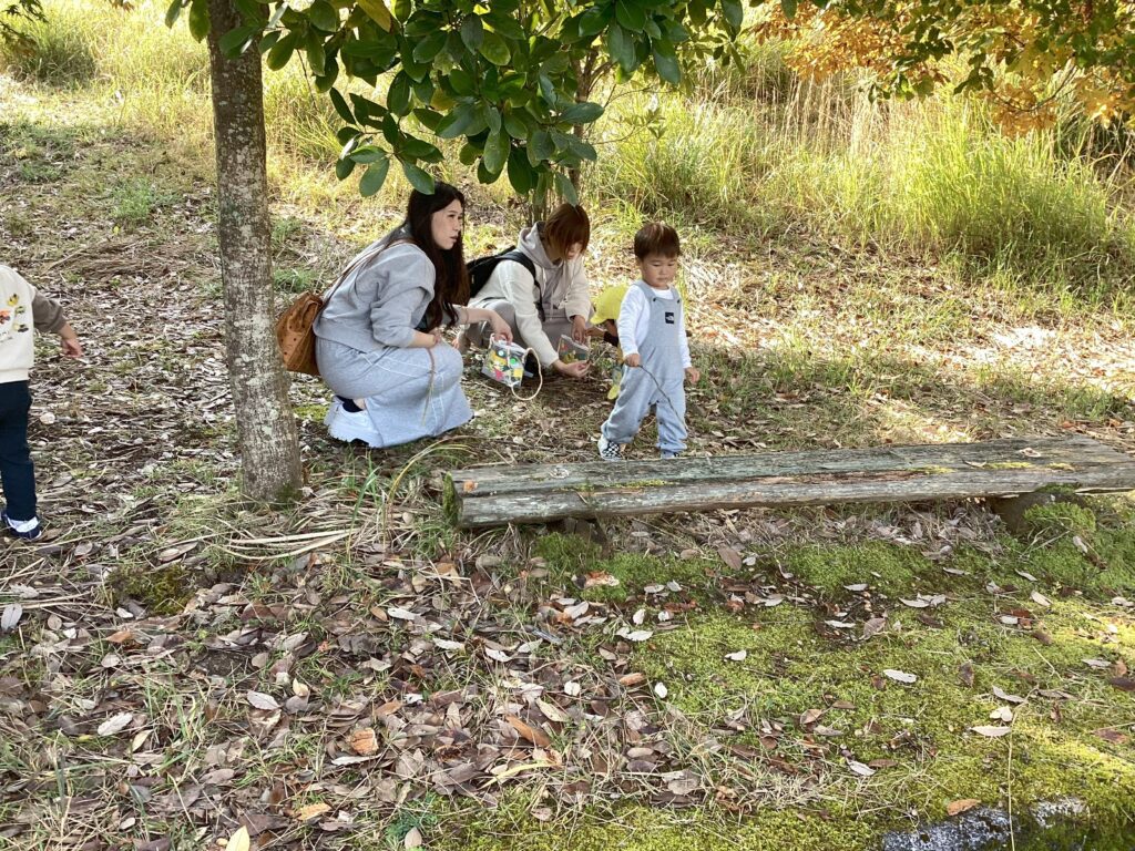 親子遠足の自然探索の様子。木の下の落ち葉の中で、ママたちがしゃがみ込み、子どもがどんぐりや木の実を探している、秋の自然体験の瞬間。