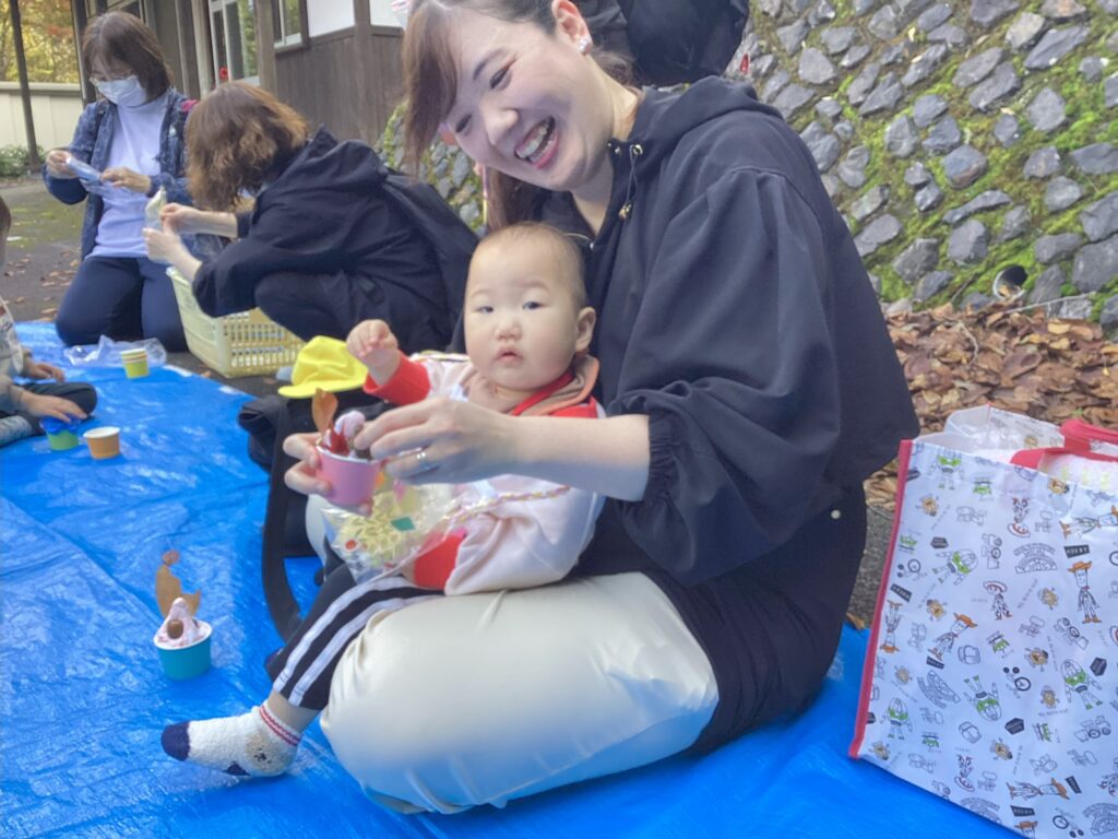 親子遠足で制作活動の様子。青いシートの上で、ママがタイムカプセル園児を抱っこしながら笑顔で、どんぐりや木の実を使ったカップケーキ制作を楽しんでいる瞬間。