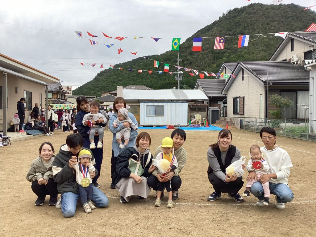 丹波市氷上のタイムカプセル保育園運動会で競技を終えた参加者（親子）の集合写真。参加者が地面にしゃがみ、万国旗が飾られた屋外グラウンドでポーズをとっている。
