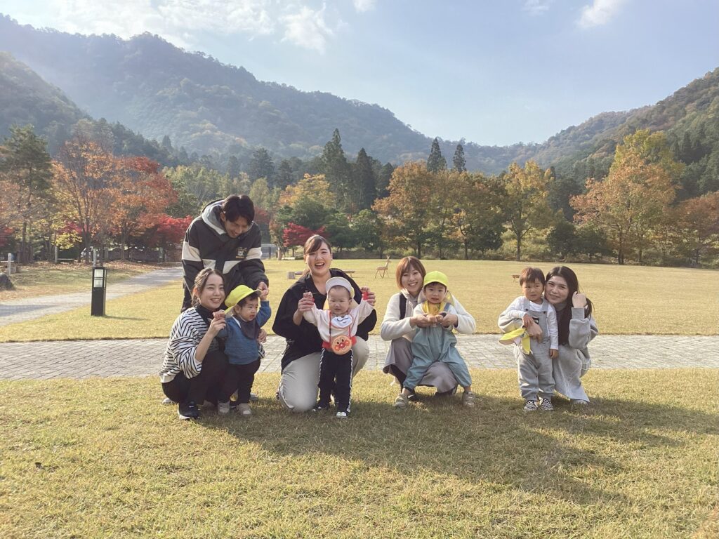 親子遠足で丹波の森公苑を背景にした集合写真。秋の紅葉が美しい山を背に、親子や先生たちが芝生の上で笑顔を浮かべている様子。