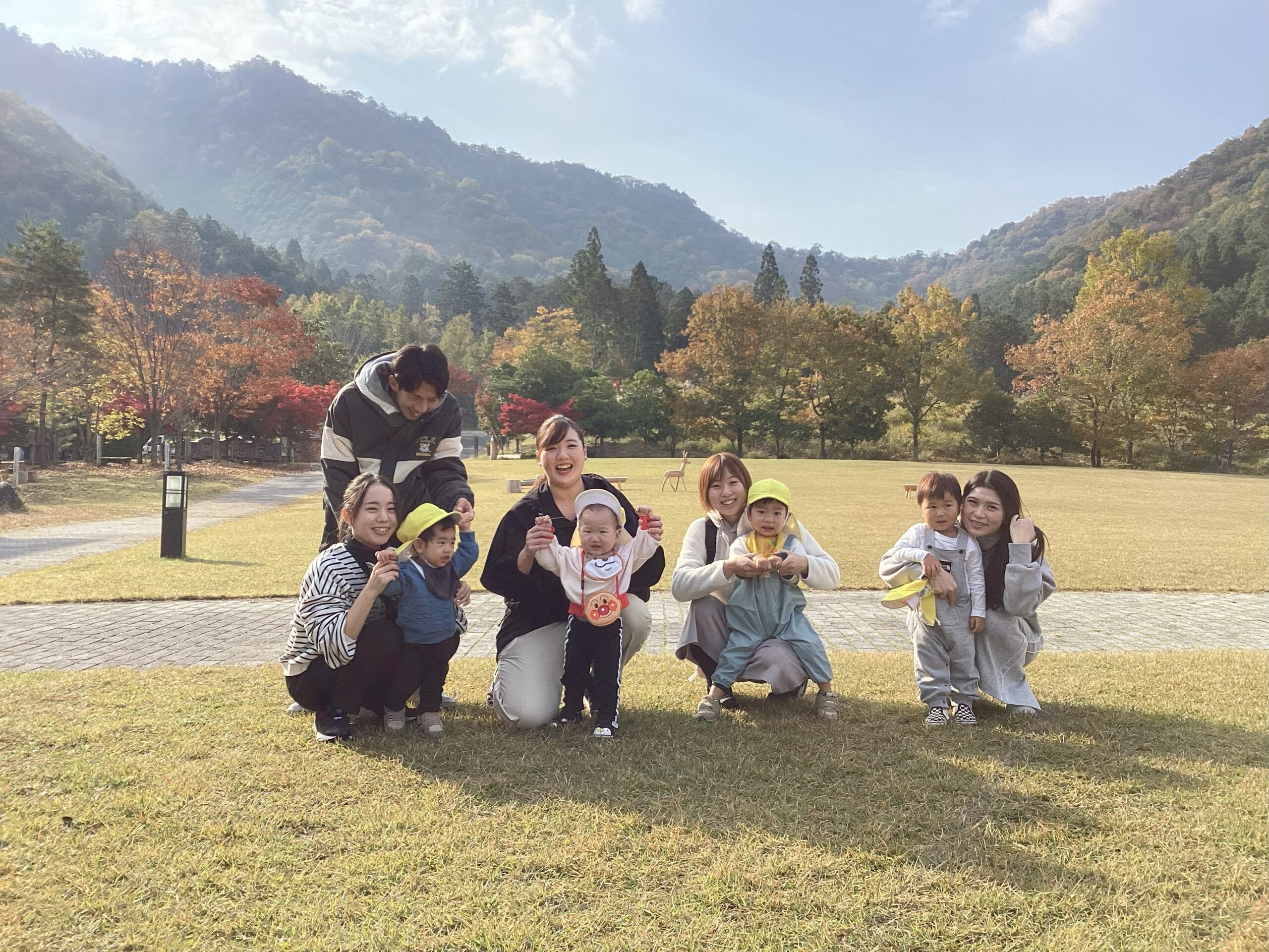 親子遠足で丹波の森公苑を背景にした集合写真。秋の紅葉が美しい山を背に、親子や先生たちが芝生の上で笑顔を浮かべている様子。