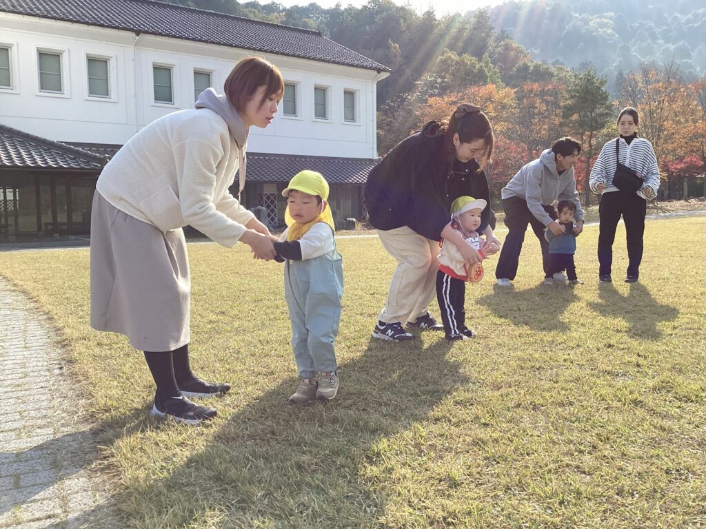 秋晴れの丹波の森公苑の芝生を歩く、親子遠足に参加したママたちとタイムカプセルの園児たち。子どもたちはデニムのオーバーオールや黄色い帽子を被り、手を繋いだり抱っこされたりして楽しそうにしています。