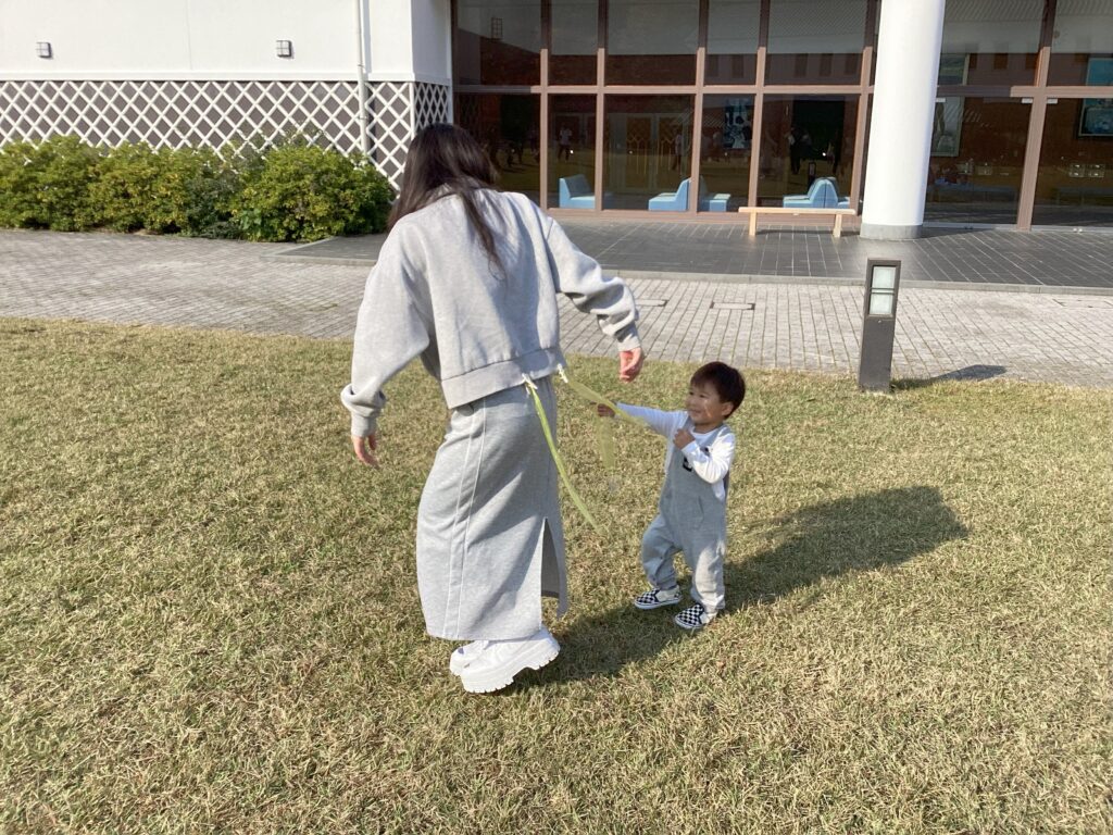 親子遠足でのしっぽとりゲーム。満面の笑顔のタイムカプセルの園児が、ママの腰についた緑色のしっぽを引っ張って捕まえようとしている、活発な遊びの瞬間。