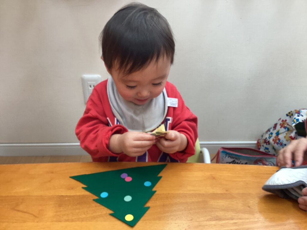 2歳児クラスの園児が、小さなシールをそっと剥がしてクリスマスツリーに貼る様子。集中した眼差しと指先の動きから、小さな成功体験を積み重ねている成長の瞬間。