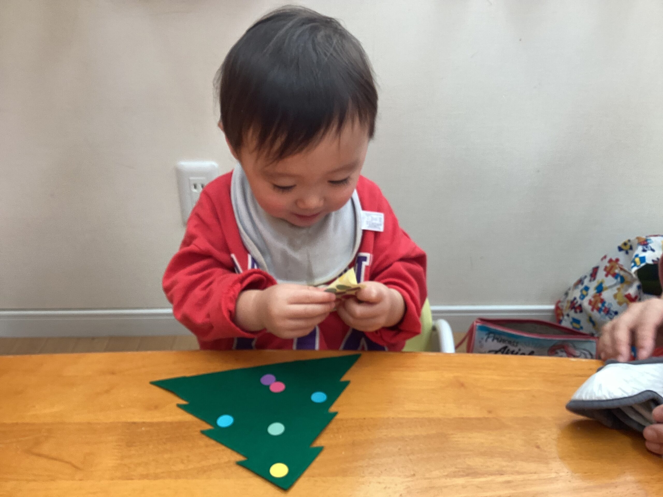 2歳児クラスの園児が、小さなシールをそっと剥がしてクリスマスツリーに貼る様子。集中した眼差しと指先の動きから、小さな成功体験を積み重ねている成長の瞬間。