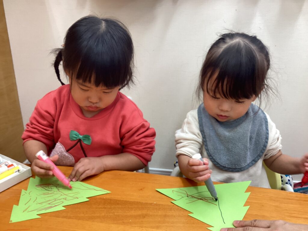 兵庫県丹波市のタイムカプセル保育園にて、2歳児クラスの子どもたちが真剣な表情でクリスマス製作に取り組む様子。緑色の画用紙で作ったツリーに、カラーペンで自由に色を塗る。