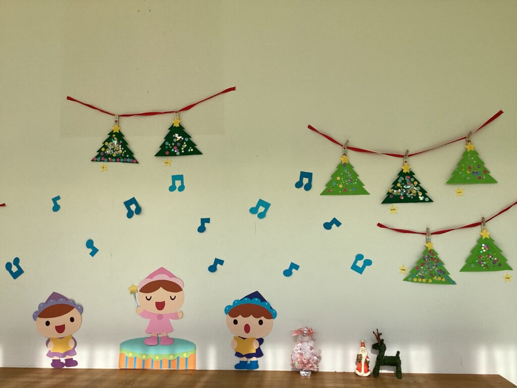 兵庫県丹波市のタイムカプセル保育園にて、2歳児クラスの子どもたちが製作したクリスマスツリーの作品が飾られた壁面。シールやスパンコールで装飾された緑色のツリーが、赤い紐に木製クリップでガーランド風に吊るされている。