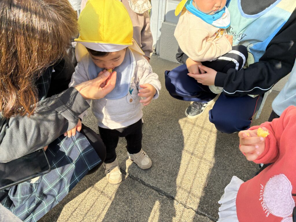 焼き立てのお芋を「あーん」と口にするお子様と、それを優しく見守る先生の眼差し。画面越しに温度感が伝わる、安心感あふれるひとコマ。