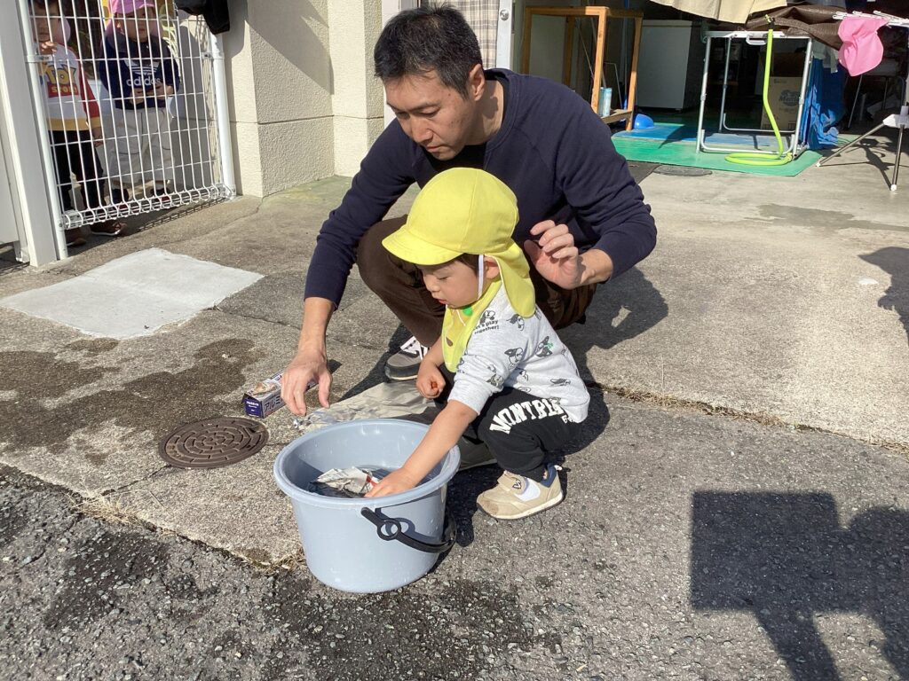 園長先生と一緒に焼き芋の下準備をする男の子。新聞紙で包んだお芋を、小さな手で一生懸命バケツの水に浸している、リトルわんぱく保育園での一コマ