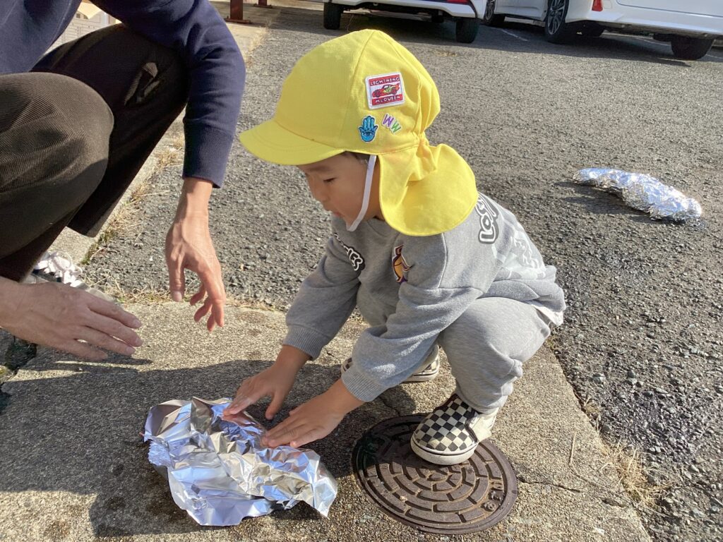 焼き芋大会の準備で、地面に座ってお芋をアルミホイルで包むタイムカプセル保育園の園児と、側でサポートする保育士
