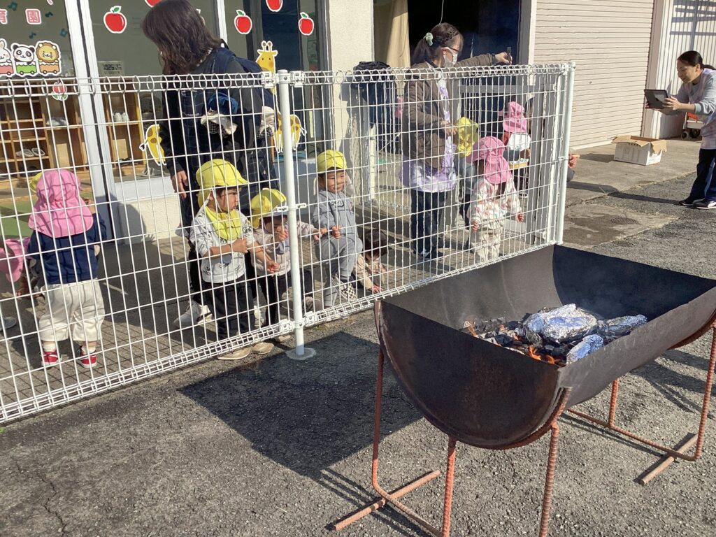 園庭に設置された焚き火台でお芋を焼く様子を、安全な距離から興味津々で見つめるタイムカプセル保育園の園児と保育士。