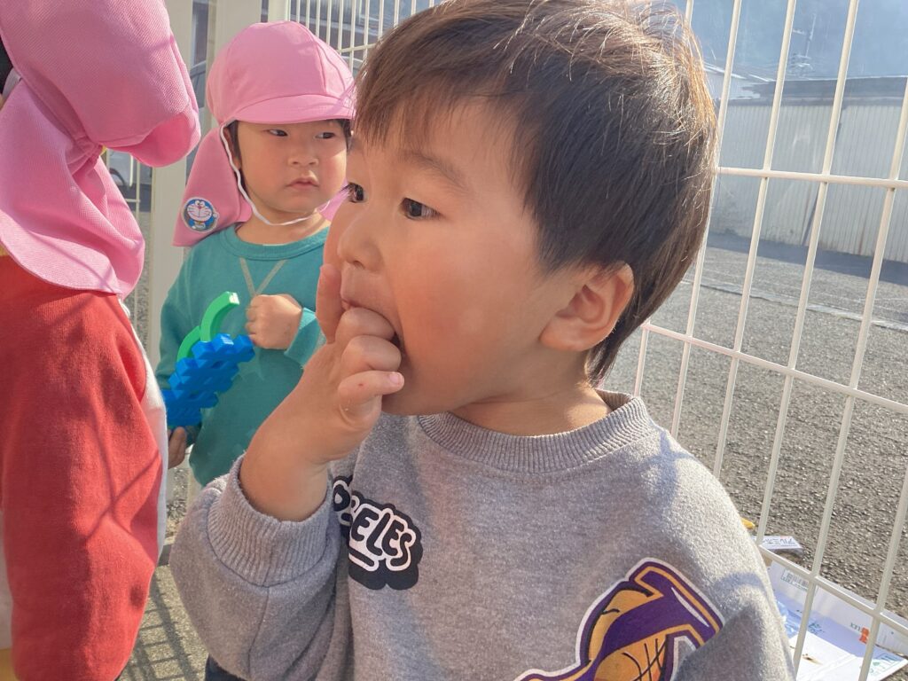 タイムカプセル保育園の食育体験。焼き立てホカホカのお芋を「おいしい！」と夢中で食べるお子様の輝く笑顔と、それを見守るお友だちのあたたかな日常風景。