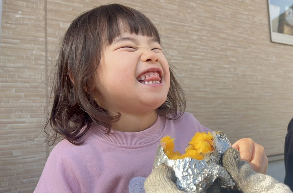 タイムカプセル保育園園児がアルミホイルに包まれたホクホクの焼き芋を大きな口を開けて食べている瞬間のクローズアップ。