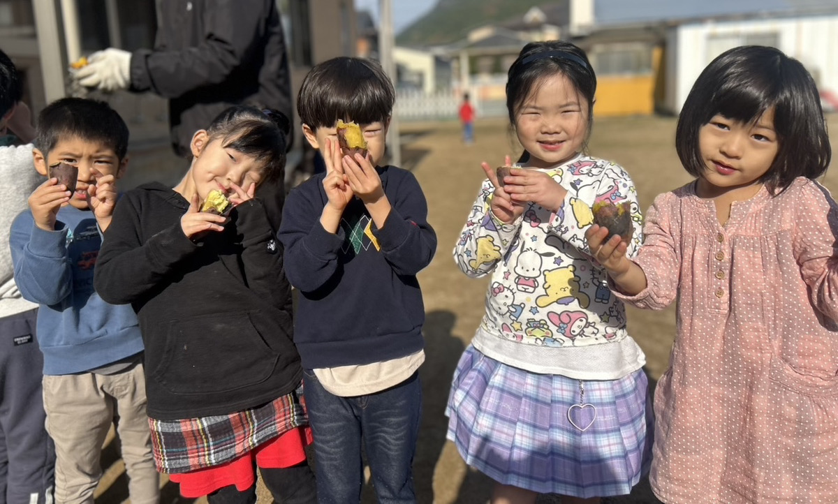 ホクホクに焼き上がったお芋を手に持ったタイムカプセル保育園の子どもたちが、満面の笑顔でカメラにポーズをとっている集合写真。喜びと達成感、そして園の賑やかで温かい雰囲気が伝わる一枚。