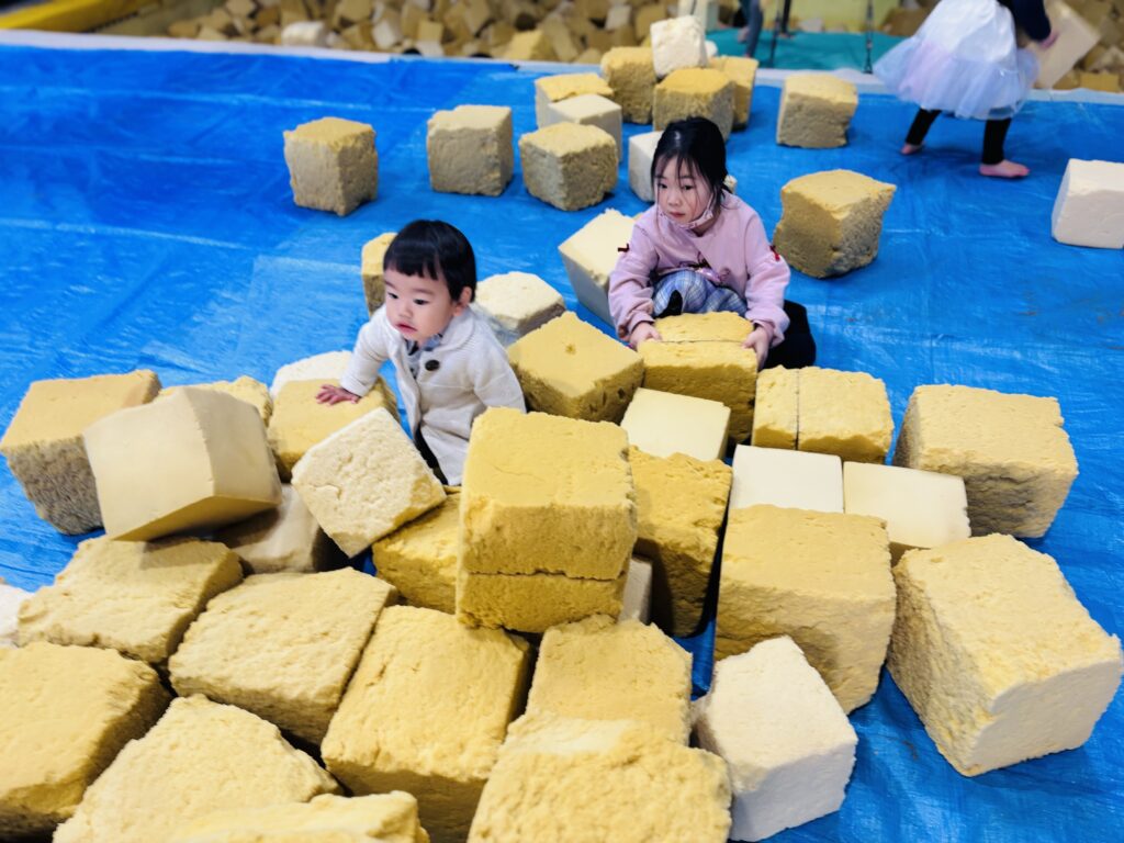 兵庫県丹波市のディナモ体操クラブ体育館で、タイムカプセル保育園の小さな子どもたち二人が協力して、青いシートの上に黄色いブロック状のスポンジを積み上げている様子。一人はしゃがみ込み、もう一人は立ってスポンジに手を添えている。