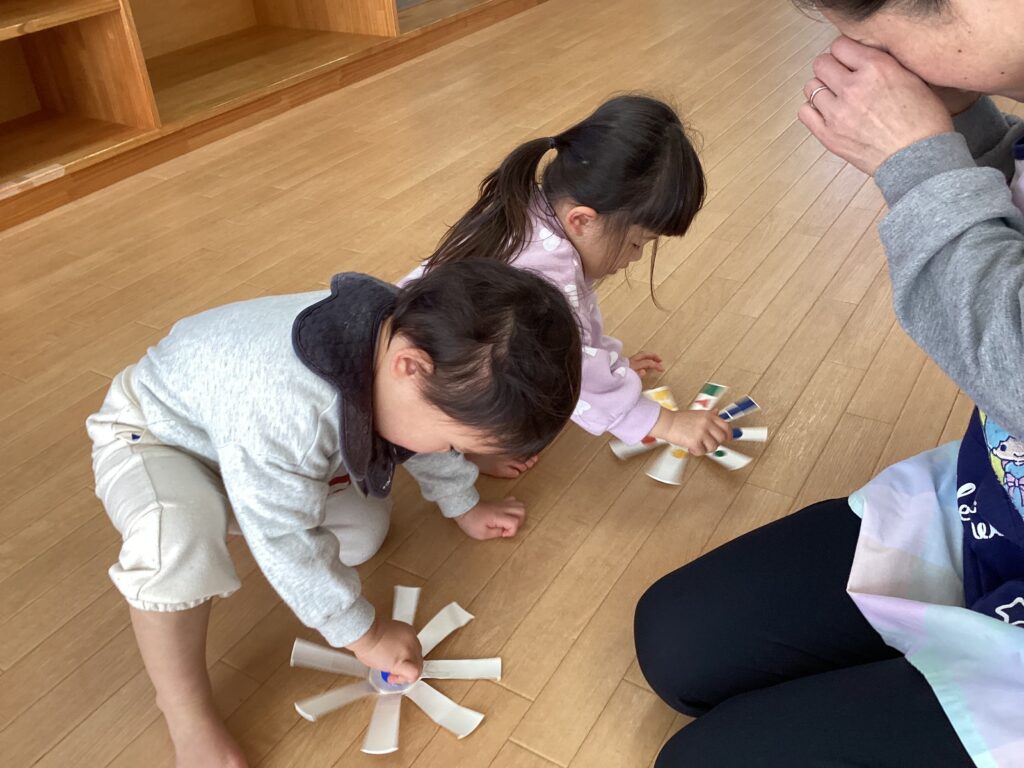 タイムカプセル保育園の室内で、自分たちで作った紙コップのコマを真剣な表情で回して遊ぶ子どもたちと、そばで見守る保育士。