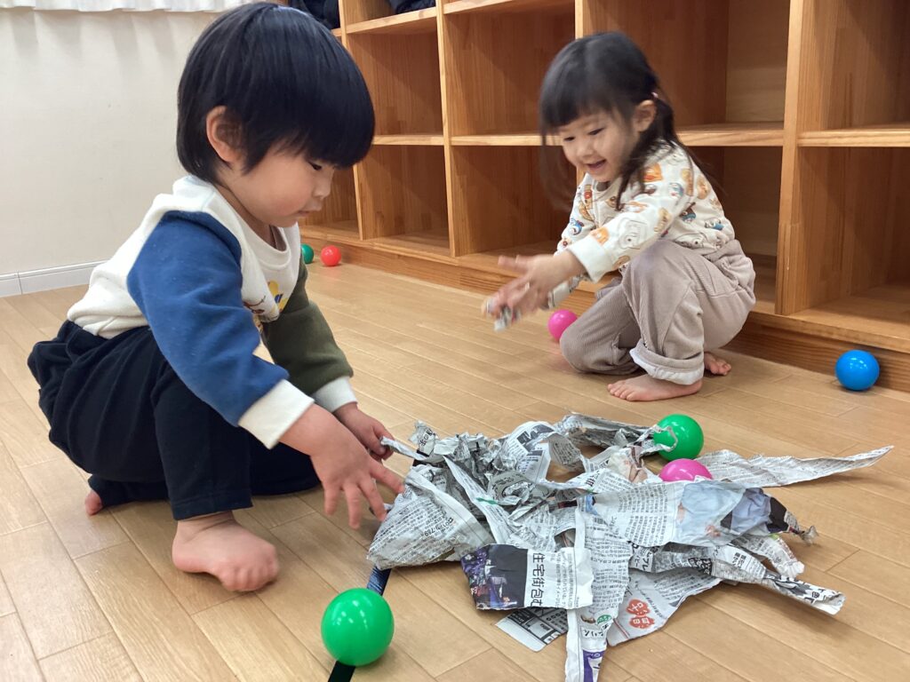 タイムカプセル保育園の床で、お友達と一緒にちぎった新聞紙を山のように集めて仲良く遊ぶ男の子と女の子