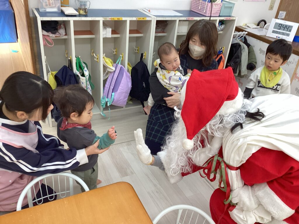 兵庫県丹波市氷上の小規模認可保育園のクリスマス会で、サンタクロースの手にタッチをして交流を楽しむ1歳児クラスの子どもの様子