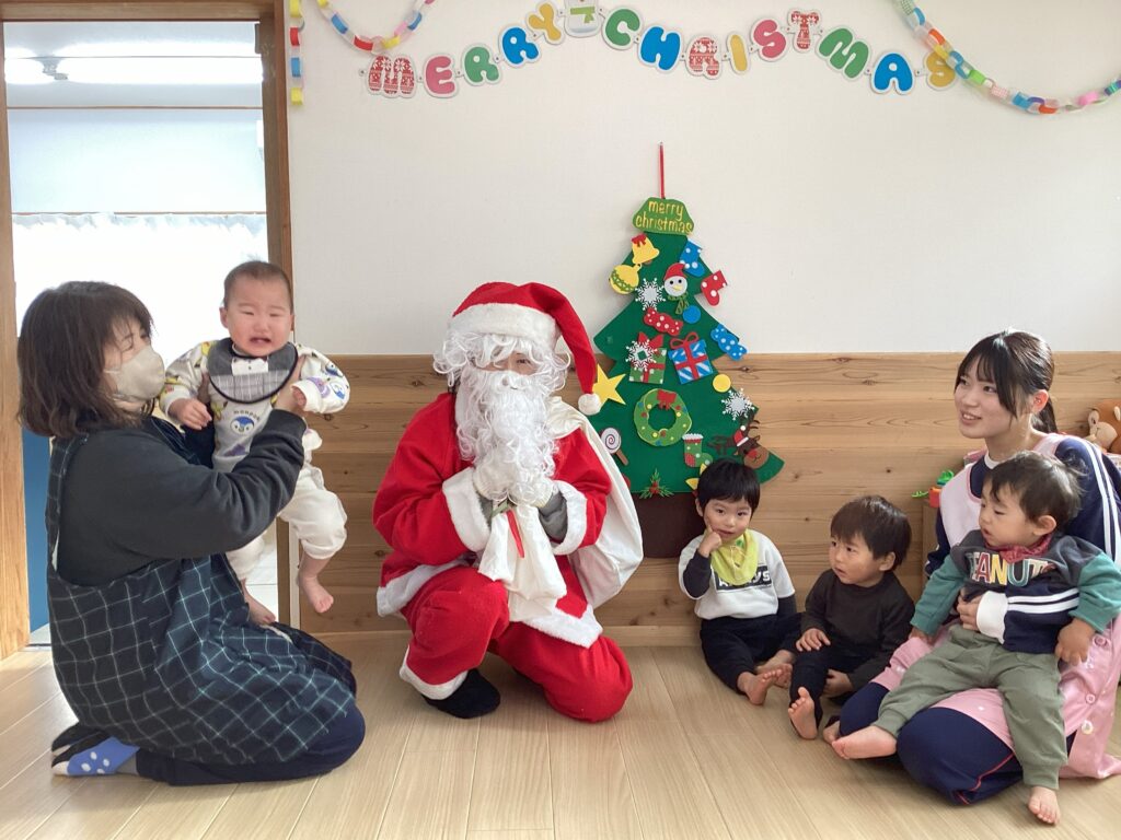 兵庫県丹波市の小規模認可保育園でのクリスマス会集合写真。サンタクロースに驚いて泣いてしまう0歳児と、笑顔を見せる1歳児、保育士たち