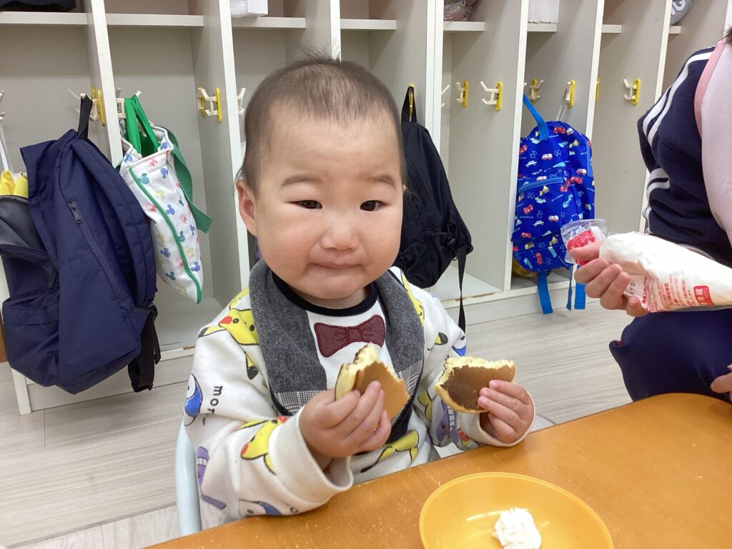兵庫県丹波市氷上の小規模認可保育園のおやつタイム。焼き上がった手作りホットケーキを両手に持ち、大きなお口で頬張る0歳児クラスの子どもの満足気な表情