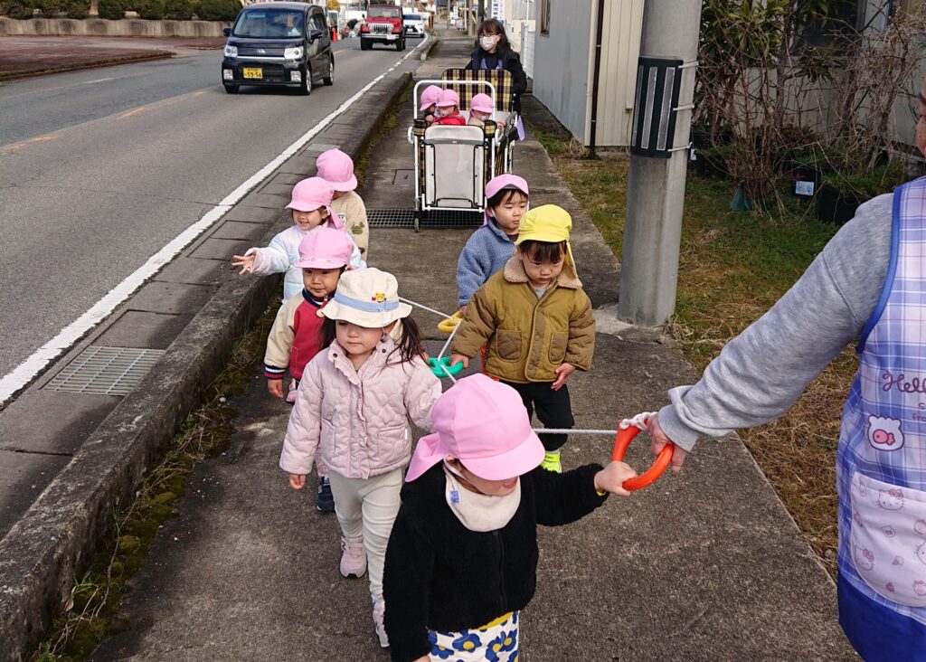 タイムカプセル保育園のお散歩風景。お散歩ロープを上手に握って公園へ向かう1・2歳児クラスの子どもたちと散歩車に乗る子どもたち