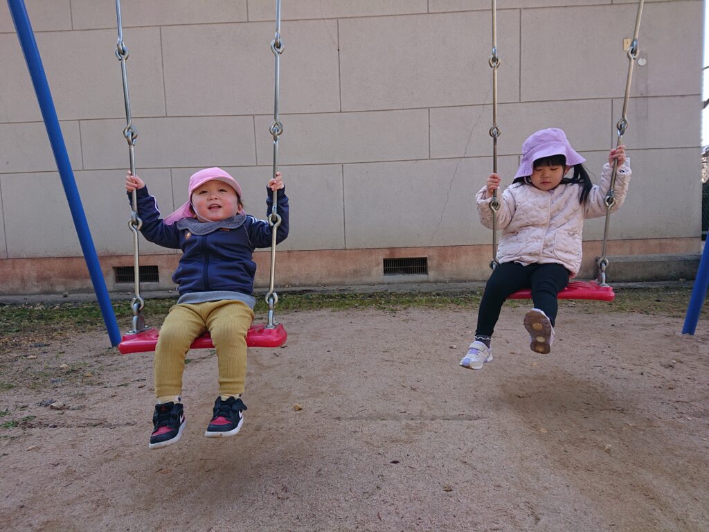 タイムカプセル保育園のお外遊び。公園のブランコに順番に乗って、笑顔でゆらゆらと揺れる1・2歳児クラスの子どもたち