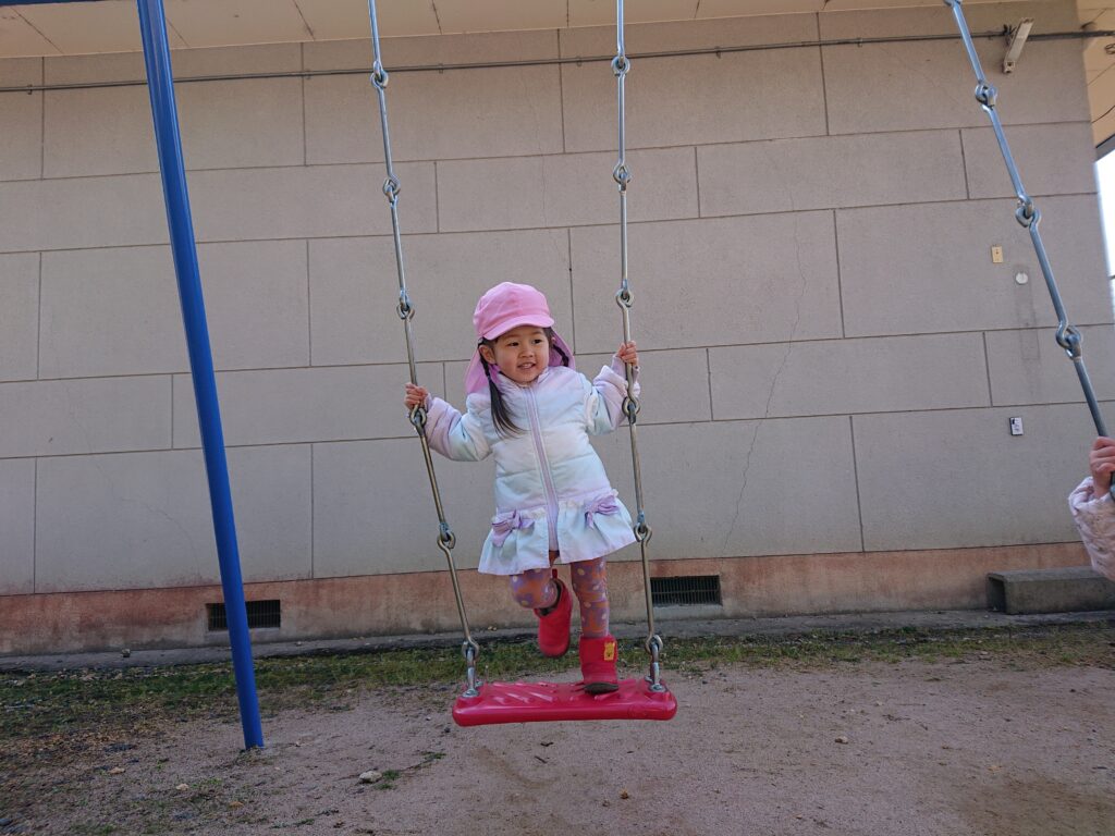 タイムカプセル保育園の公園遊び。ブランコで上手に「立ちこぎ」に挑戦し、風を切って笑顔を見せる2歳児クラスの女の子