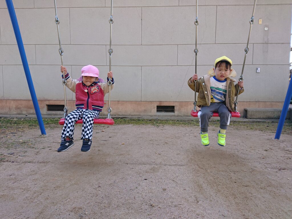 タイムカプセル保育園の公園遊び。ブランコに順番に座って、しっかりとチェーンを握りながら安全に遊ぶ1・2歳児の子どもたち