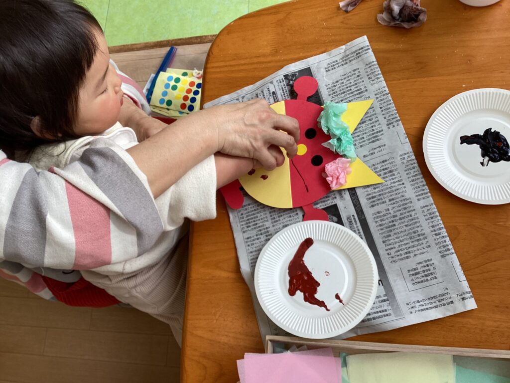 兵庫県丹波市のリトルわんぱく保育園での節分制作。保育士と一緒に鬼のお面に花紙を貼る子どもの様子