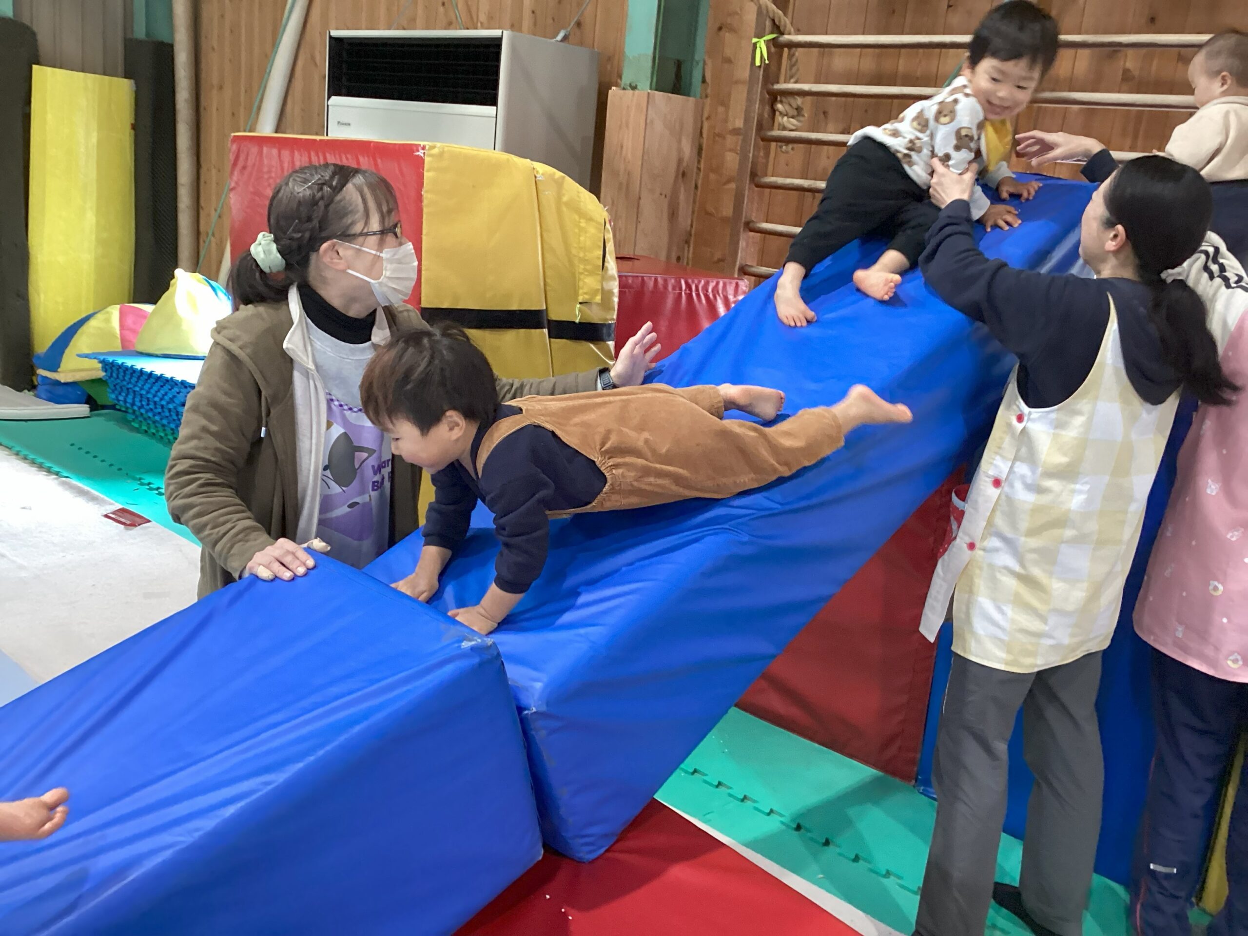 タイムカプセル保育園の体操あそびの様子。保育士のサポートのもとマットの滑り台で遊ぶ子ども