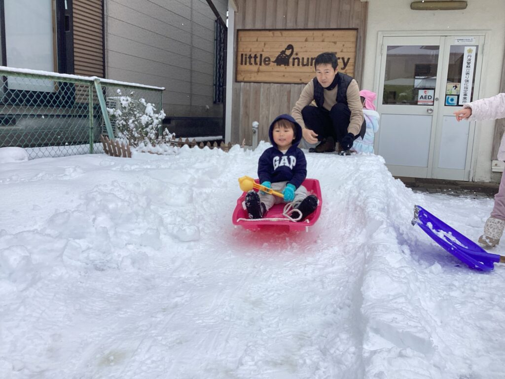 兵庫県丹波市氷上のタイムカプセル保育園での雪遊び。園長先生が見守る中、手作りの雪の滑り台をピンクのそりで滑る子どもの様子